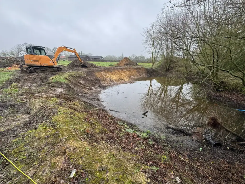 Excavator digging beside rural pond on overcast day